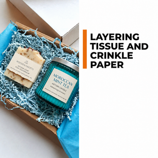 Blue tissue lining and pale blue crinkle paper around artisan soap and a teal candle in a kraft gift box.