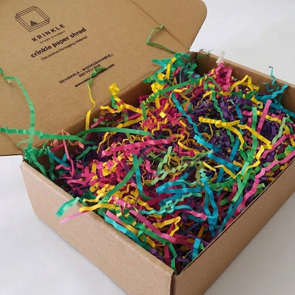 An open kraft cardboard box filled with bright multicolored shredded paper filler, photographed on a plain white background. The inside lid shows the printed brand name 'Kreinkle' and product text.