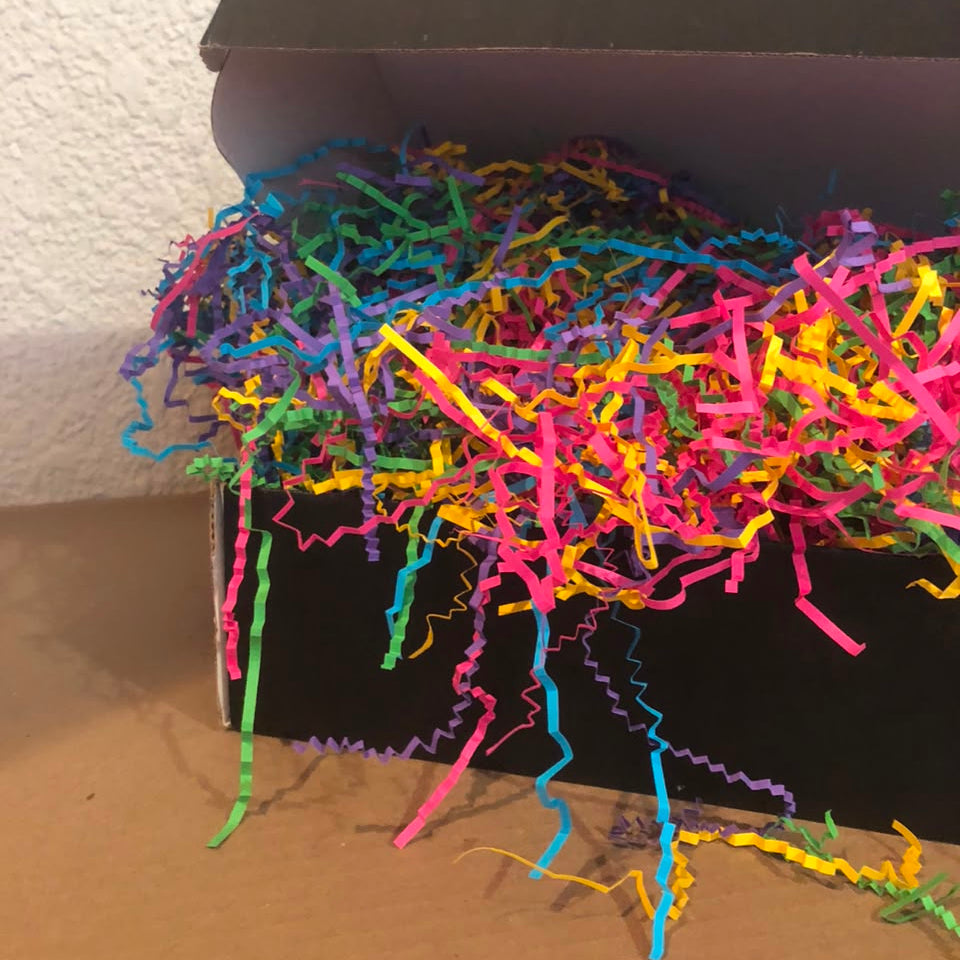 A black cardboard box filled with bright multicolored shredded paper filler, photographed indoors against a textured off-white wall and tan floor.