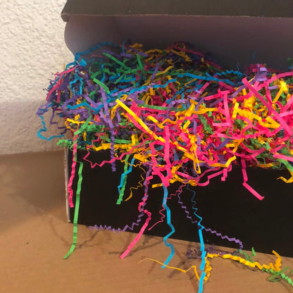 A black cardboard box filled with bright multicolored shredded paper filler, photographed indoors against a textured off-white wall and tan floor.